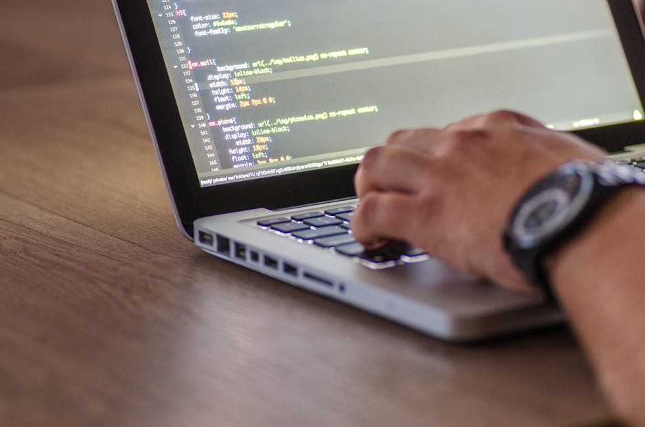 A close-up image of a person's hand with a wristwatch, typing on a silver laptop placed on a wooden desk. The screen displays lines of code with colorful syntax highlighting. The background is blurred
