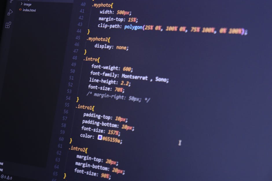 A close-up image of a person's hand with a wristwatch, typing on a silver laptop placed on a wooden desk. The screen displays lines of code with colorful syntax highlighting. The background is blurred